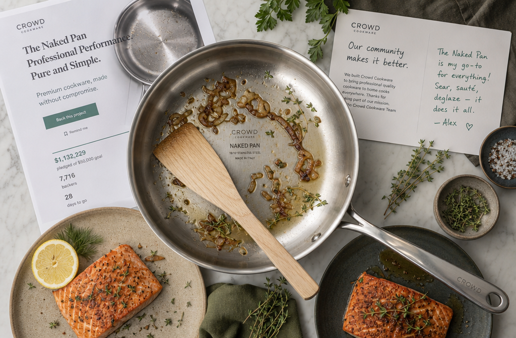 Crowd Cookware Review 2026 Landscape format (1536x1024) overhead flat-lay photograph of a Crowd Cookware stainless steel Naked Pan on a marble kitchen surface surrounded by fresh ingredients — seared salmon, caramelized onions, herbs — with a wooden spatula resting across the pan. Background includes a printed Kickstarter campaign page and a community feedback card. Soft natural window light from upper left, muted earth tones with steel and green accents, food editorial photography style, high resolution, magazine quality.