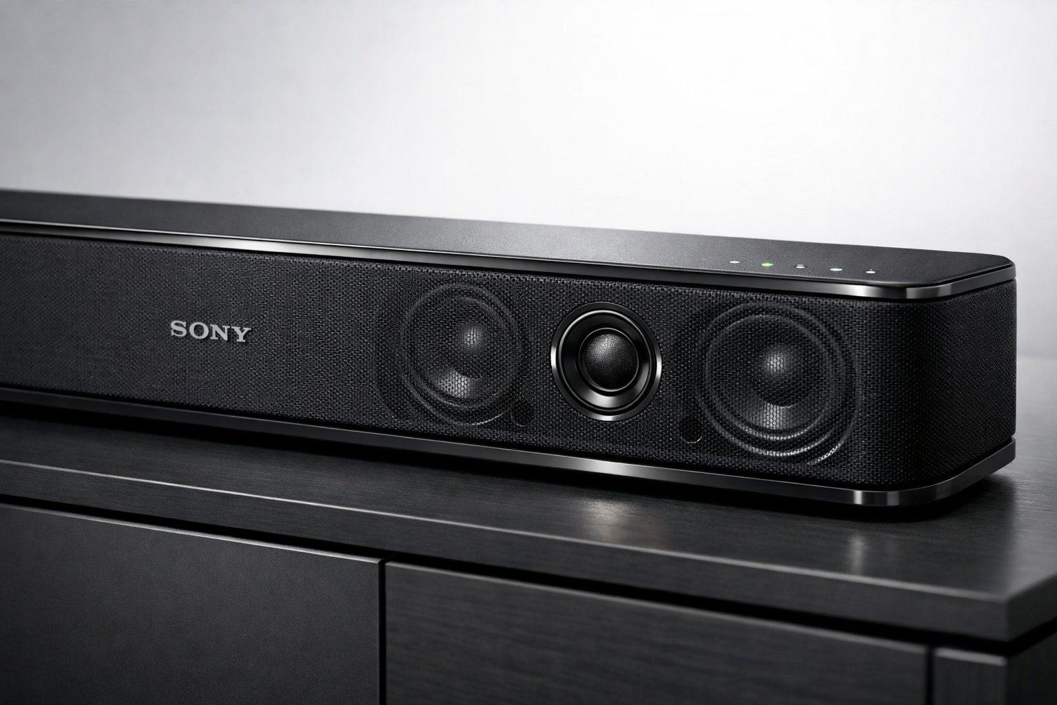 Sony Bravia Theatre Bar 5 Detailed landscape format (1536x1024) image showing close-up product photography of Sony Bravia Theatre Bar 5 soundbar from front three-quarter angle, highlighting premium build quality with matte black finish, fabric grille texture clearly visible, subtle Sony branding, LED indicators, multiple speaker drivers visible through mesh, metallic accents catching light, positioned on modern entertainment console with soft studio lighting, clean white background with subtle gradient, professional product photography style with sharp details and depth of field effect.