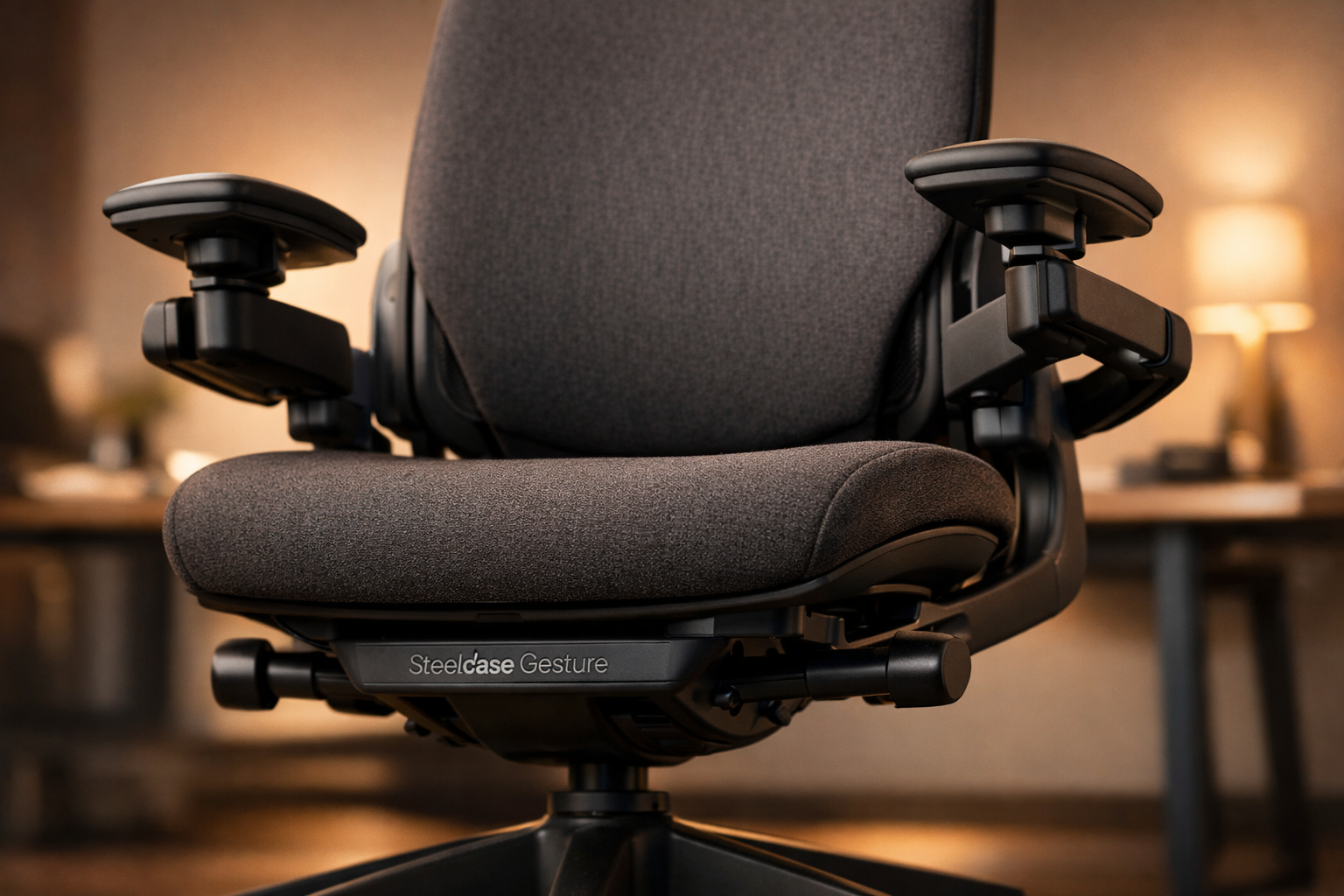 Herman Miller Aeron vs Steelcase Gesture Steelcase Gesture chair photographed from a low front angle emphasizing its signature 360-degree adjustable arms in mid-position, upholstered seat and 3D LiveBack system visible, warm studio lighting on charcoal fabric, 'Steelcase Gesture' nameplate subtly visible on the base, modern office desk in soft background bokeh.