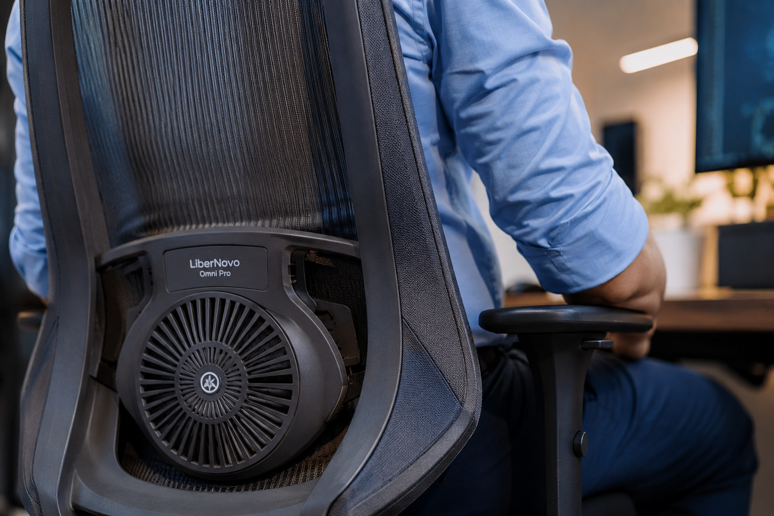 Landscape format (1536x1024) close-up editorial photograph of the LiberNovo Omni Pro chair backrest mesh and lumbar support mechanism from a low 45-degree angle, showing ventilation fan grille detail and upholstery texture. A professional seated in business casual attire demonstrates natural spinal alignment. Warm office lighting, shallow depth of field, ergonomic comfort focus, photojournalistic style with subtle blue and gray color grading. The 2026 Lineup: Omni SE, Omni Pro, and Omni Maxis