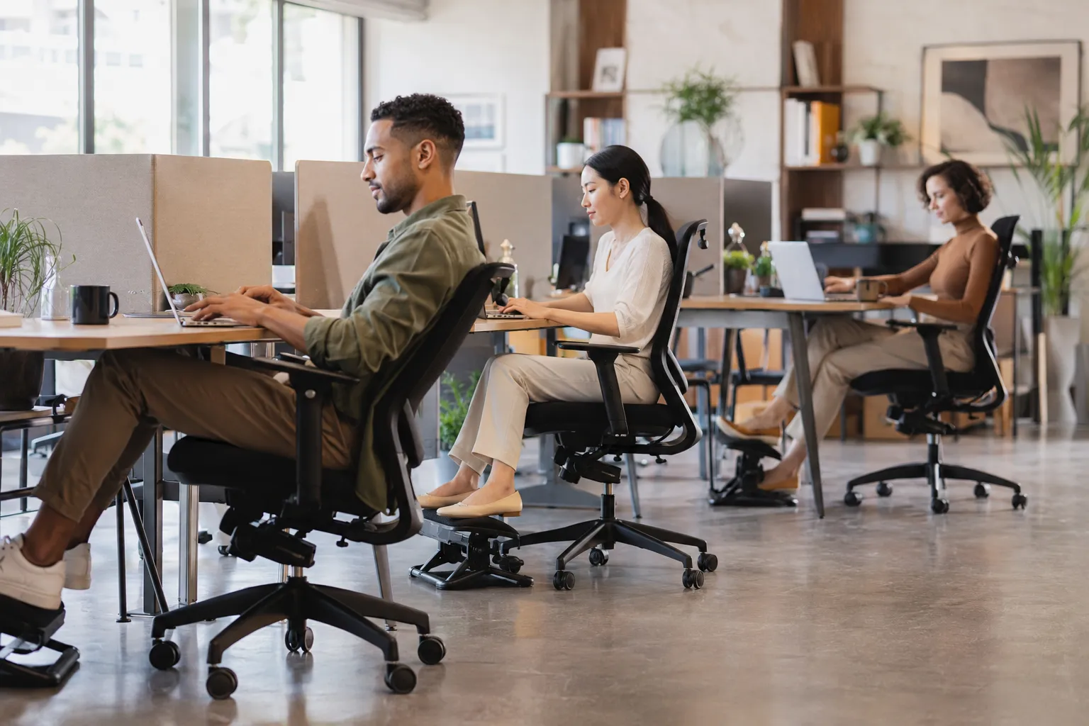 Editorial landscape photograph (1536x1024) showing a diverse group of three office workers — a tall man, a short woman, and a person of average height — each seated in ergonomic chairs at separate desks in a bright open-plan modern office. Natural daylight from large windows, warm neutral tones, each person shown in a relaxed productive posture with footrests deployed. Shallow depth of field on the foreground figure, magazine-quality composition, diverse ethnicities, professional editorial photography style, no text overlays.

Common Mistakes When Setting Up an Ergonomic Chair with Footrest