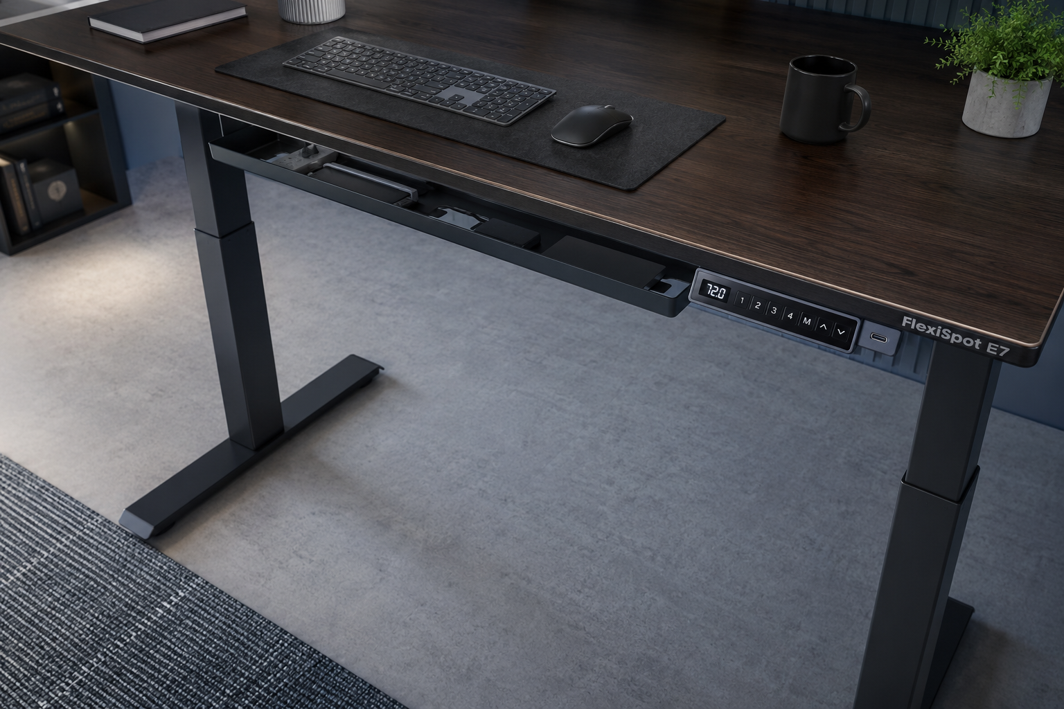 Overhead angled view of FlexiSpot E7 standing desk showing its premium LCD keypad with USB-C port and height presets, upright motor columns, and built-in cable tray underneath a wide desktop, 'FlexiSpot E7' label visible, cool modern office lighting FlexiSpot Premium Standing Desk E7