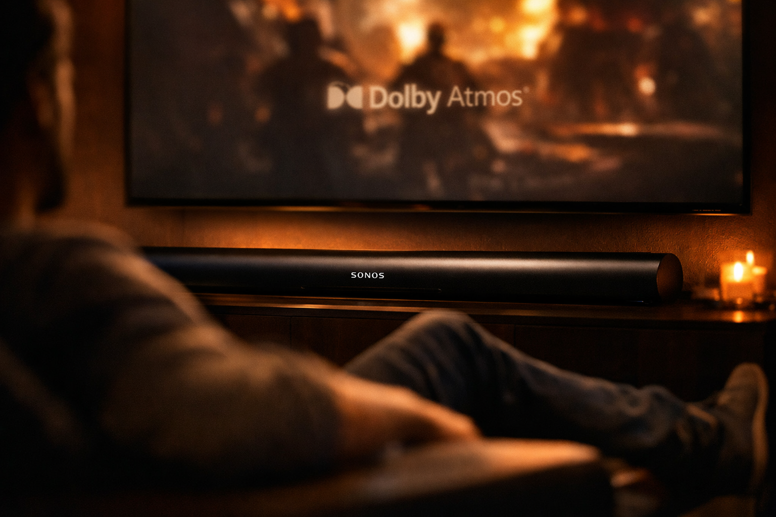 Close-up lifestyle photograph of a person sitting on a couch in a dimly lit home theater room, a Sonos Arc soundbar glowing softly beneath a large screen showing a movie, Dolby Atmos logo subtly visible on the TV display, warm cinematic lighting, shallow depth of field focusing on the soundbar, 1536x1024 landscape format, editorial home entertainment photography style, rich dark tones with amber highlights.